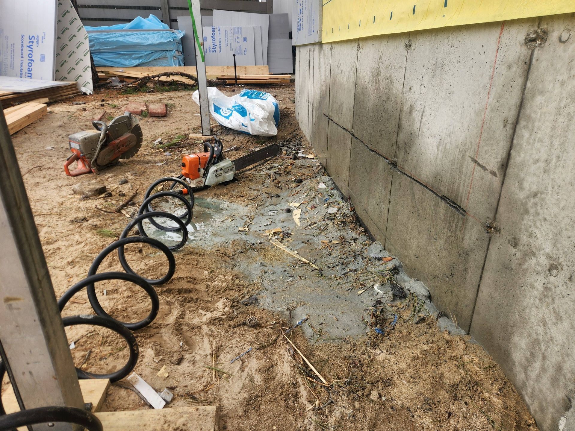 A concrete foundation wall sits on a dirt construction site with a chainsaw and coiled hose in the foreground.