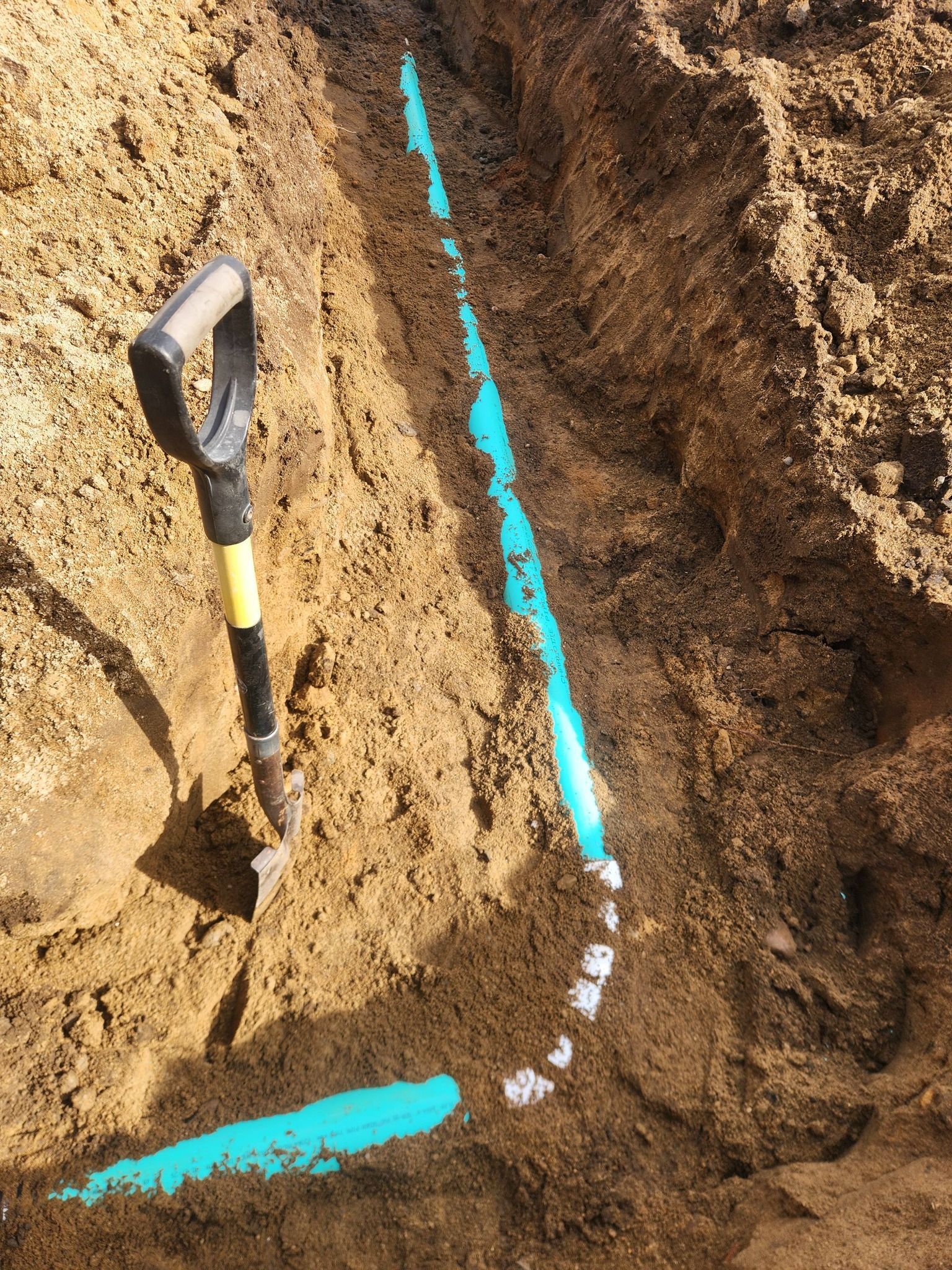 A shovel stands upright in a dirt trench next to a section of blue piping that curves through the soil.