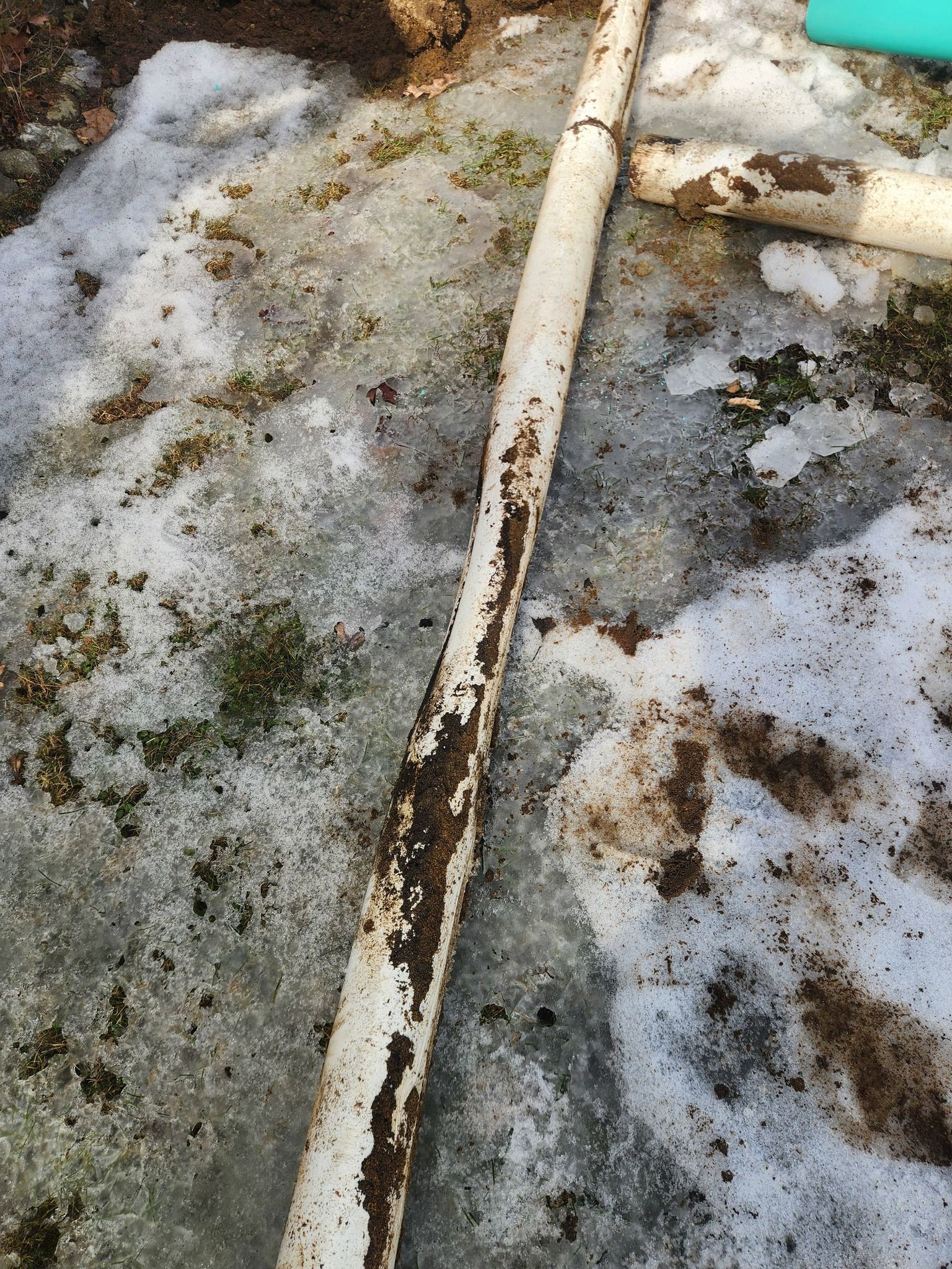 A long, white, dirt-stained PVC pipe lying on a patch of melting, slushy snow and gravel.