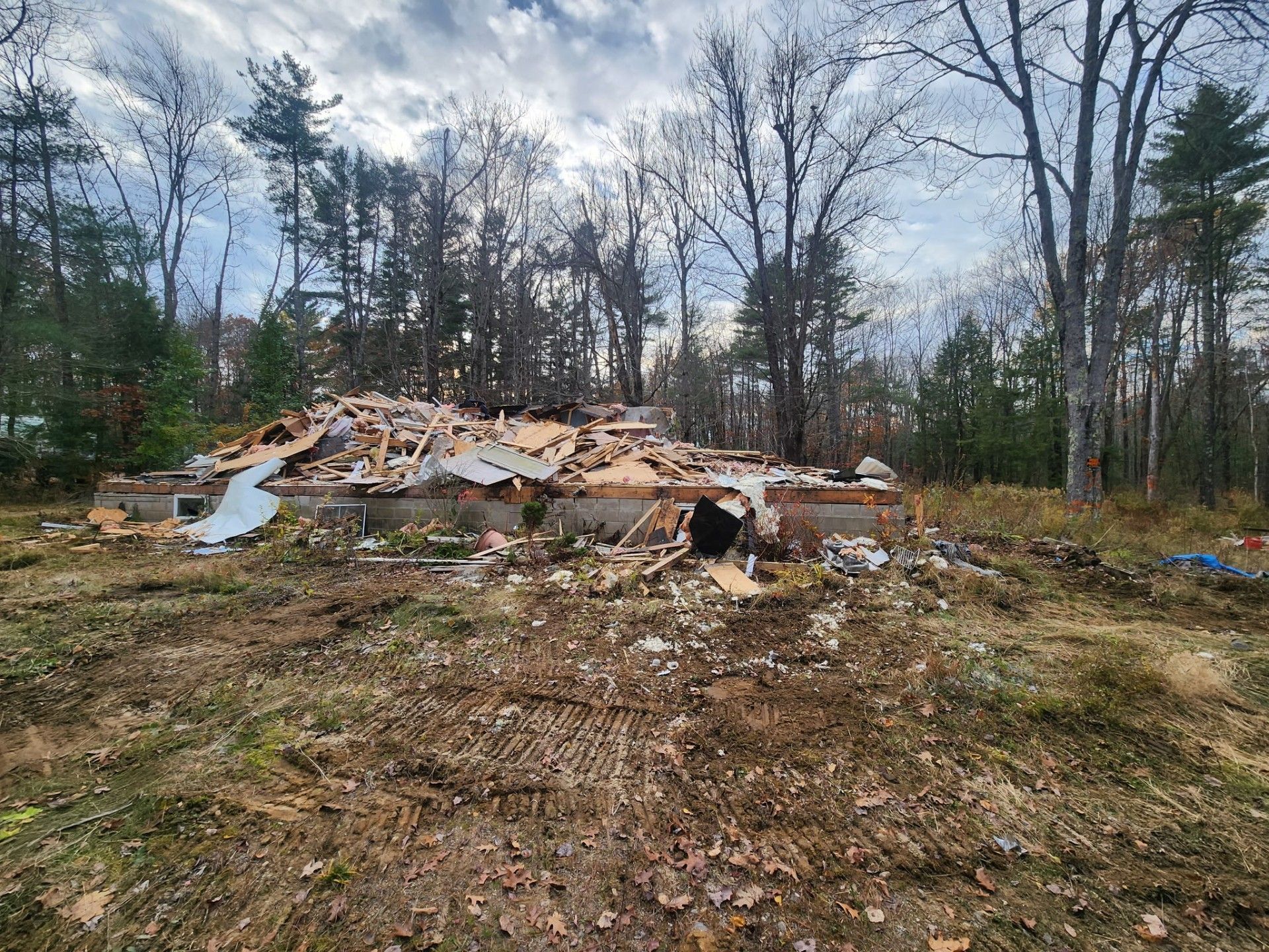 Debris of a demolished building on a brown field, with trees in the background under a cloudy sky.