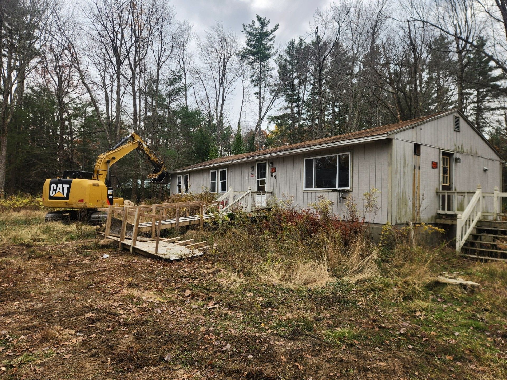 A yellow excavator is demolishing a white building with a ramp, in a wooded area.