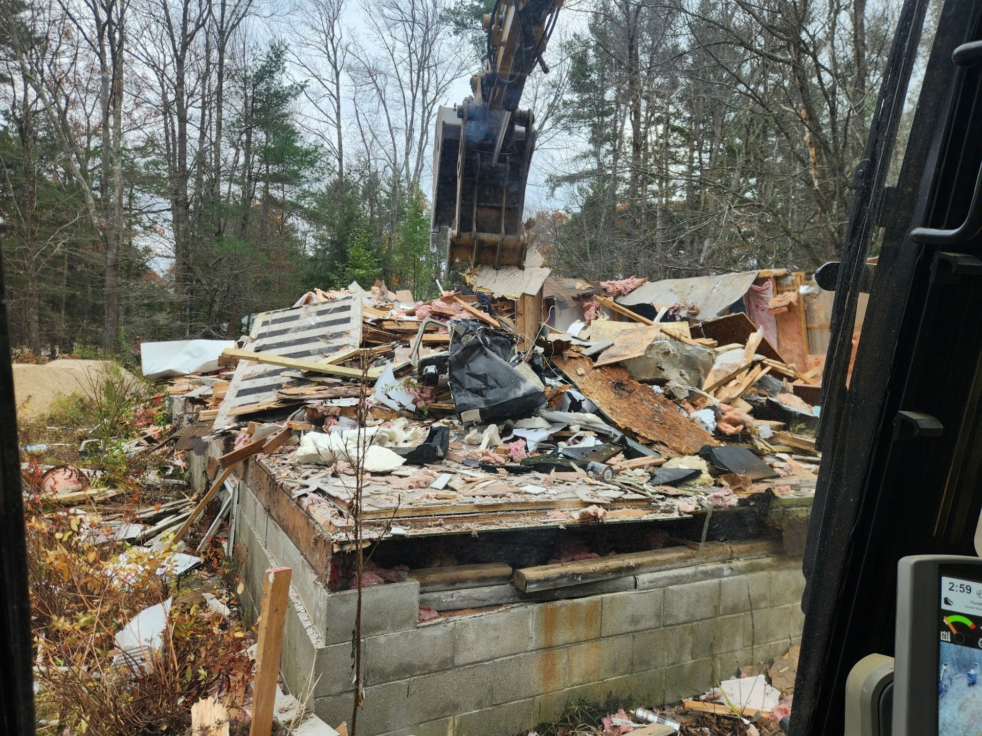 An excavator demolishes a building, showing exposed foundation and debris in a wooded area.