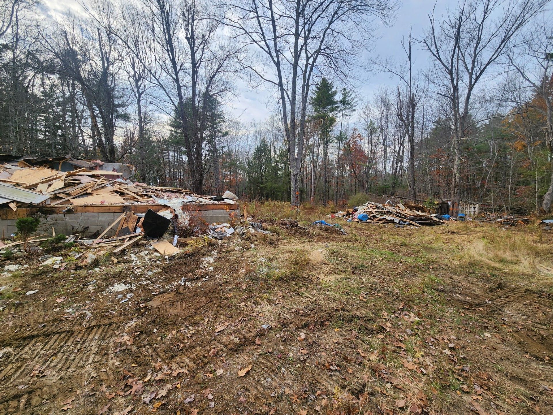 Debris and remnants of a building sit in a clearing with bare trees and a cloudy sky.