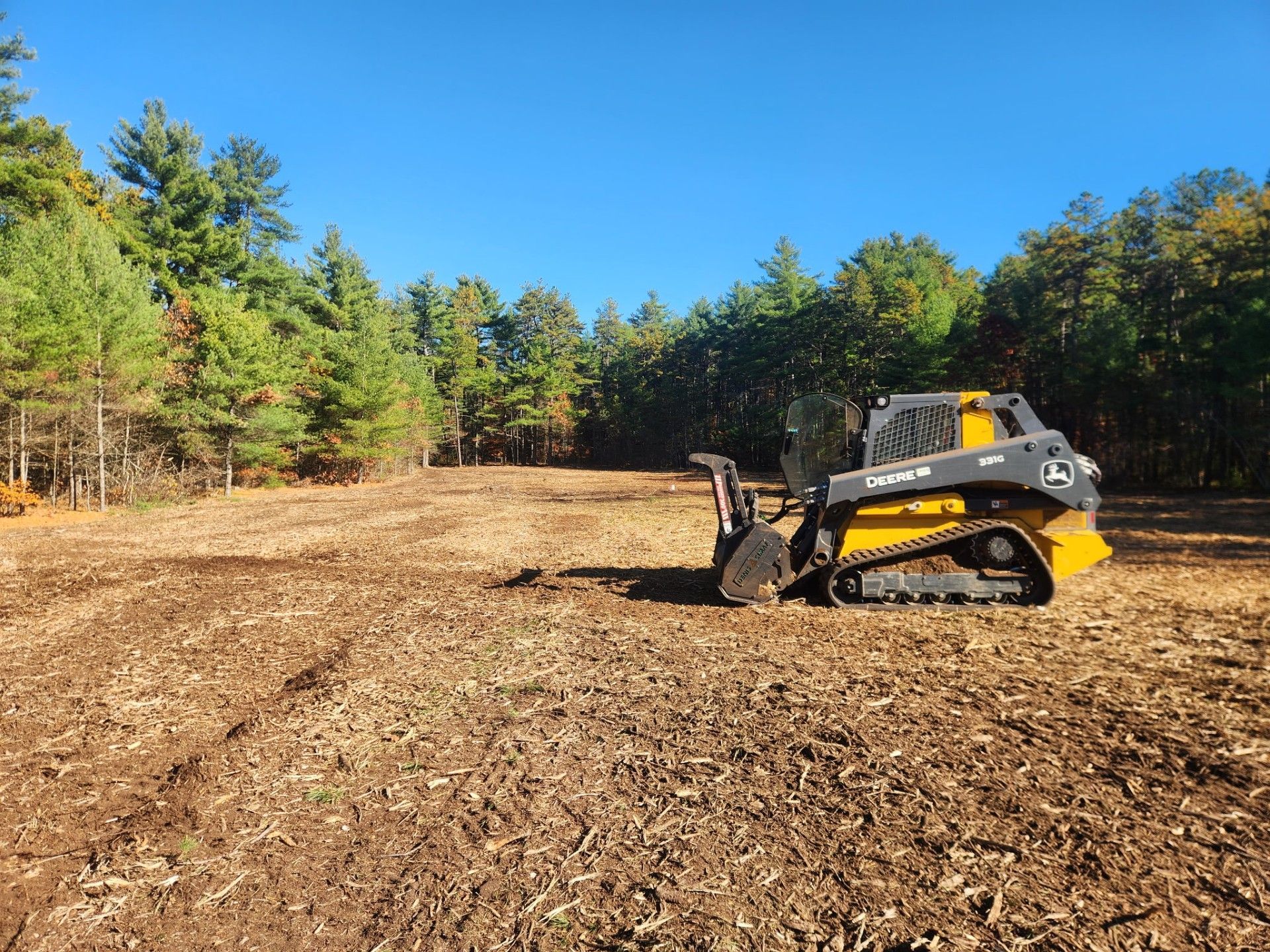 A yellow skid steer tractor on freshly cleared land, trees in the background, blue sky.