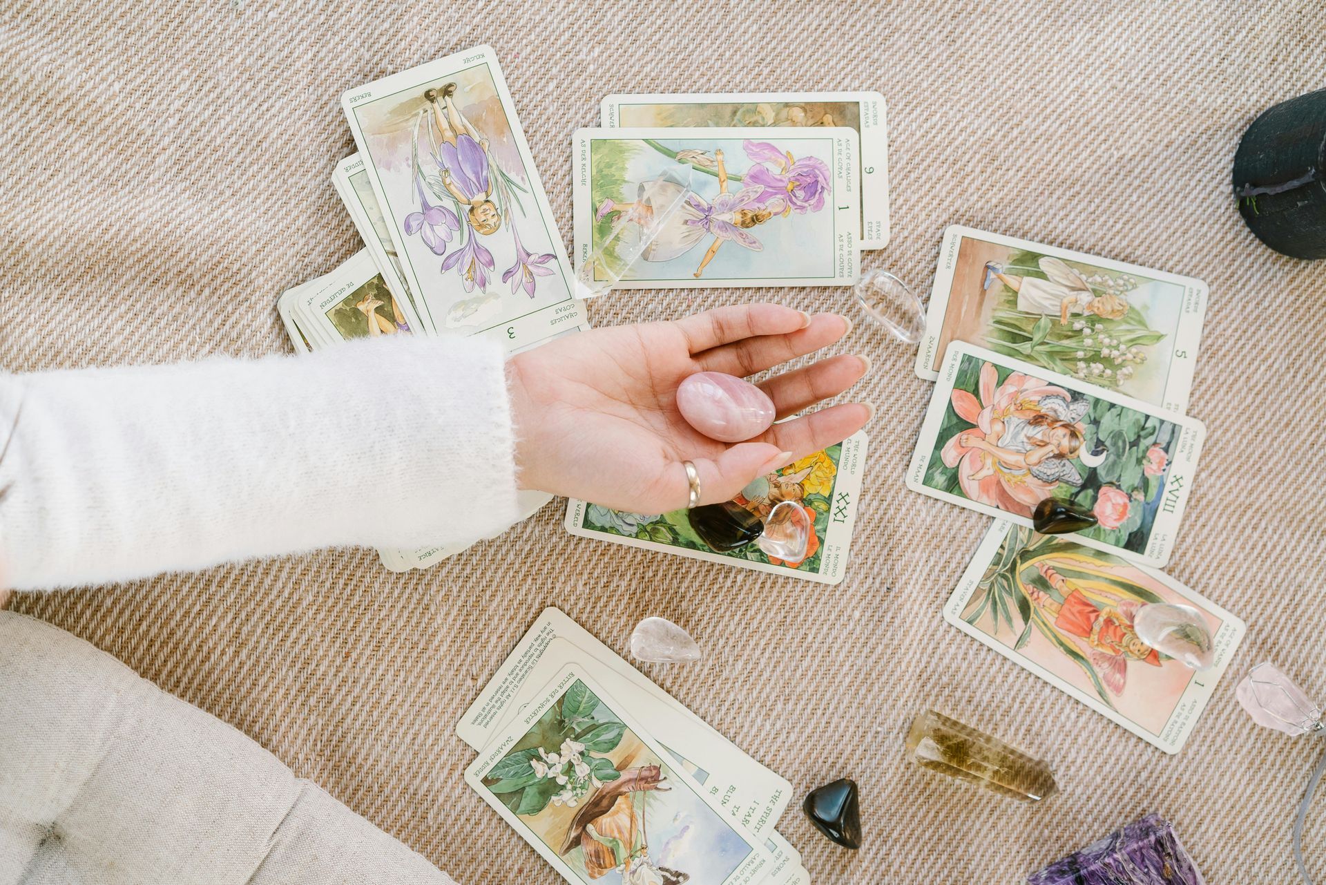 A person is holding a pink crystal in their hand surrounded by tarot cards and crystals.