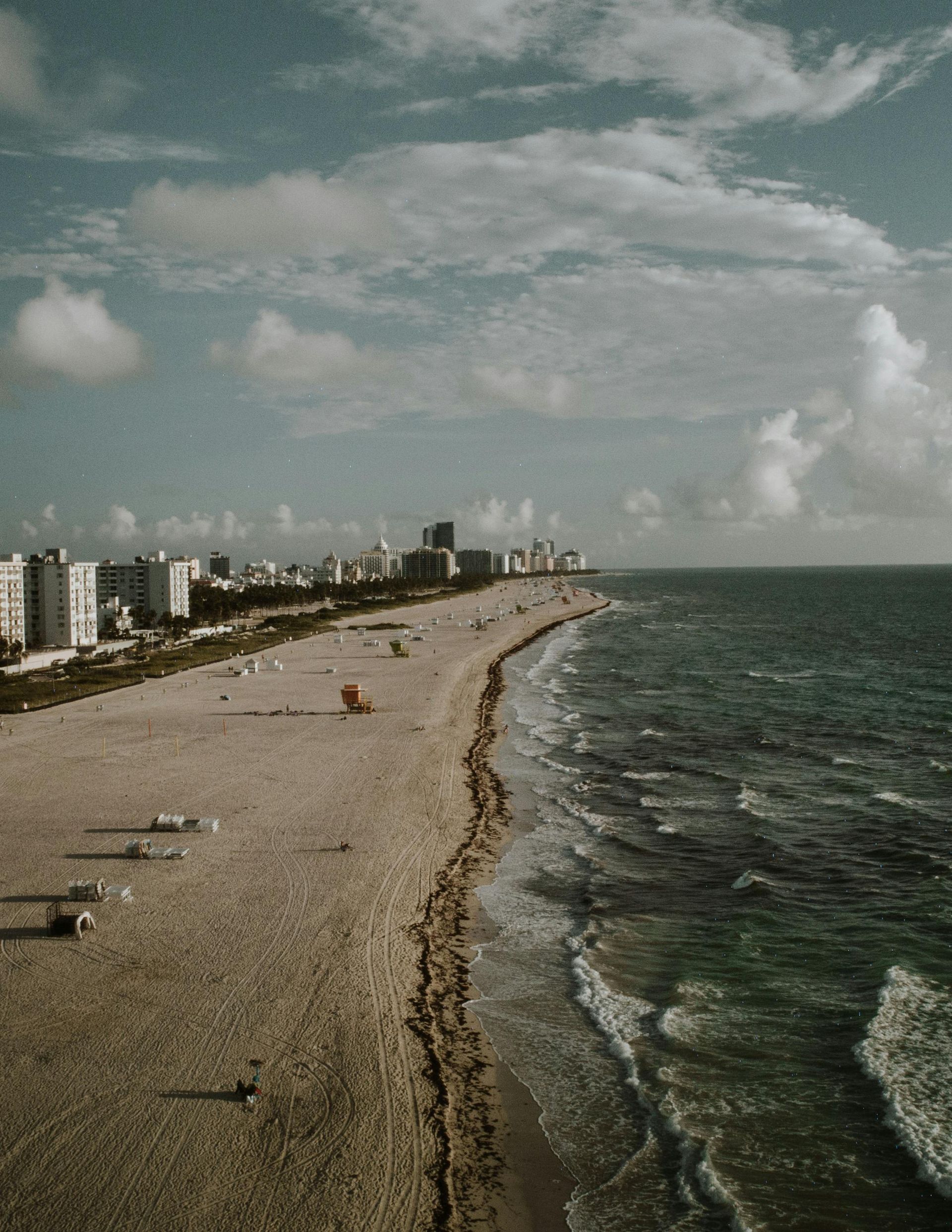A rocky beach curves along the coast beside a city skyline under a cloudy sky.