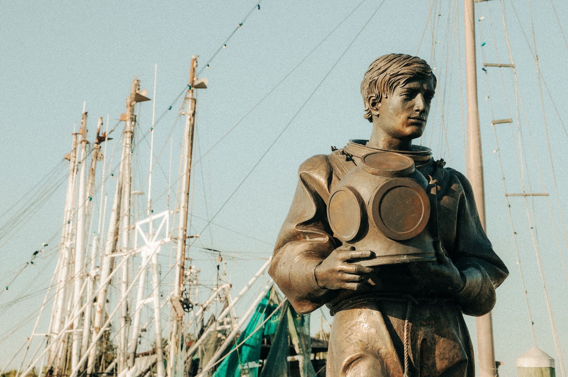 Bronze statue of a diver holding a helmet, with masts of boats in the background.