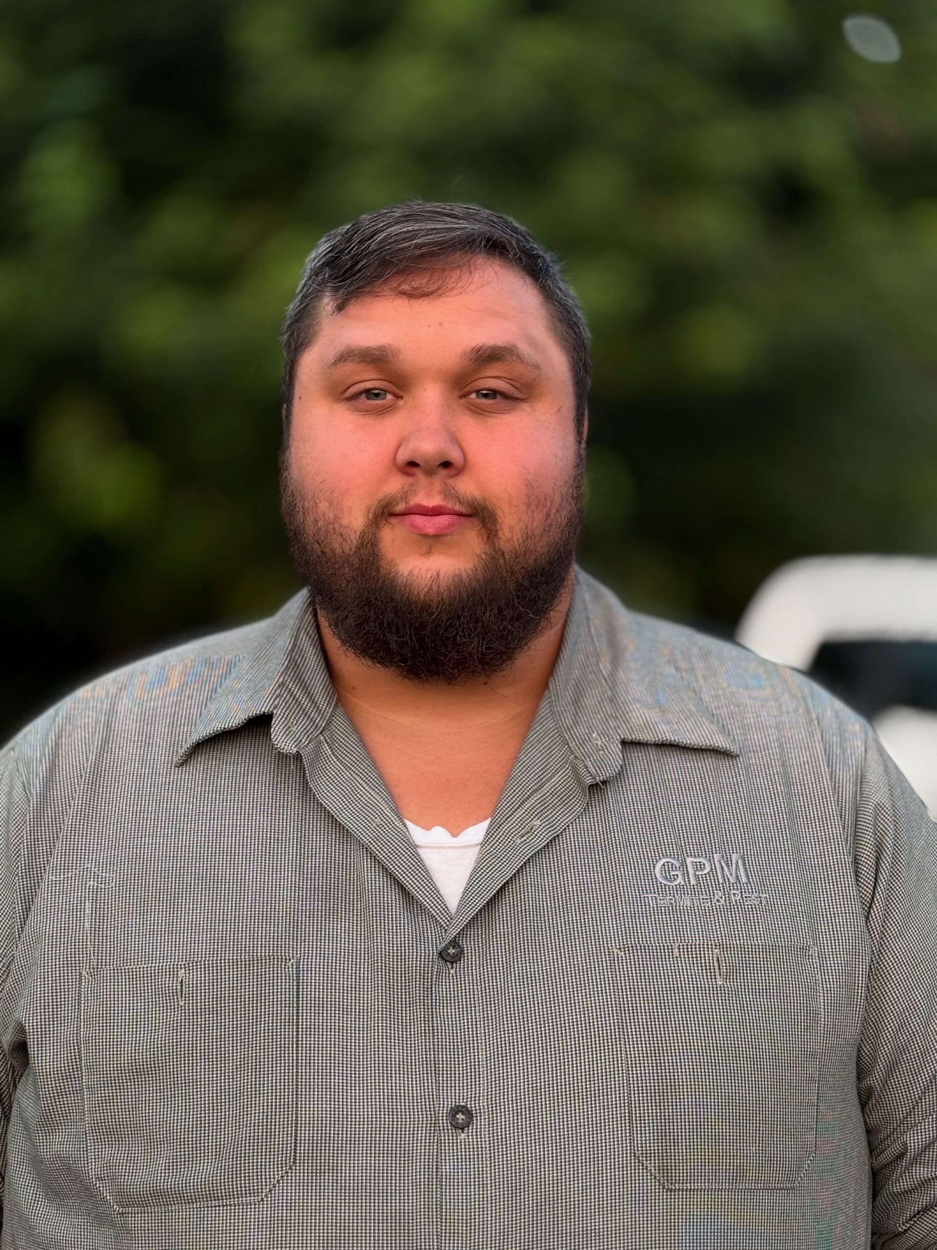 Man with brown hair and beard wearing a striped shirt, looking forward with a neutral expression. Outdoors.