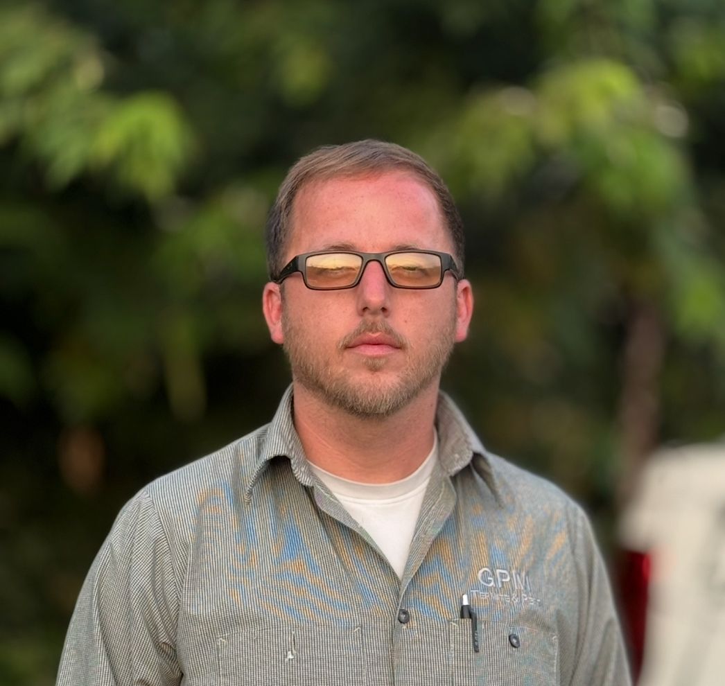 A man wearing sunglasses and a striped shirt is standing in front of trees.