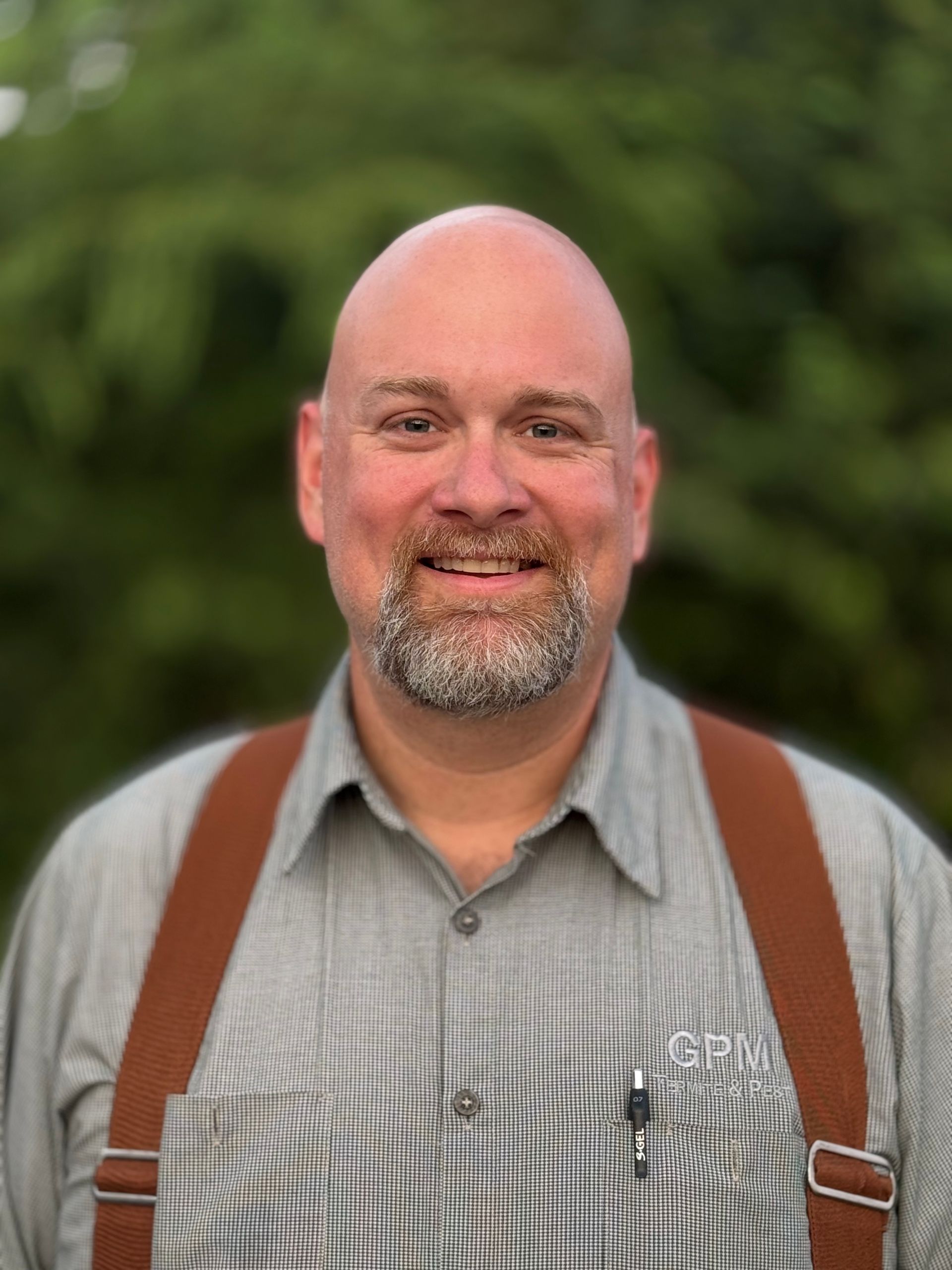 A bald man with a beard and suspenders is smiling for the camera.