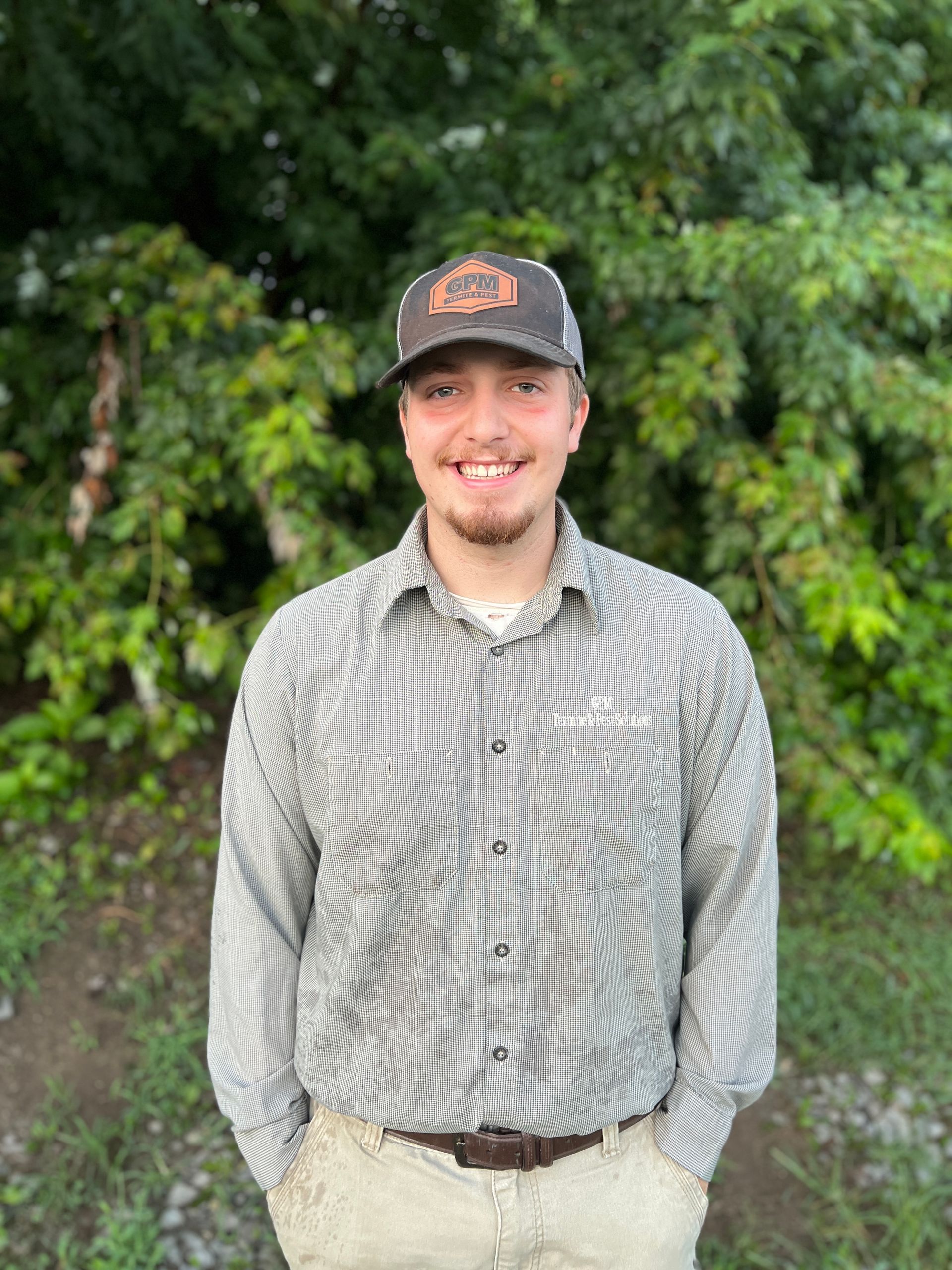 A young man wearing a hat and a striped shirt is standing in front of trees.