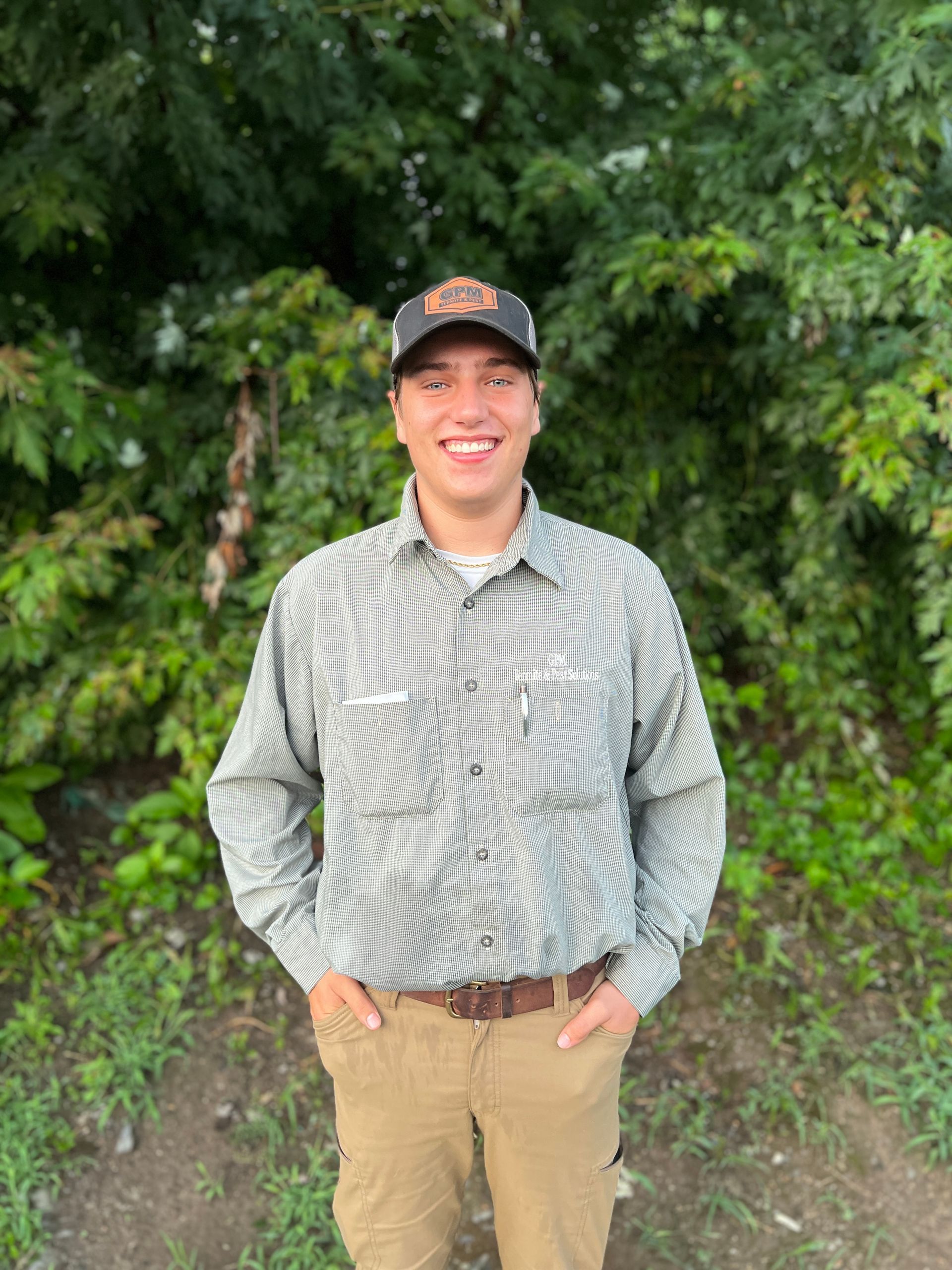 A young man wearing a hat and a shirt is standing in front of a forest.
