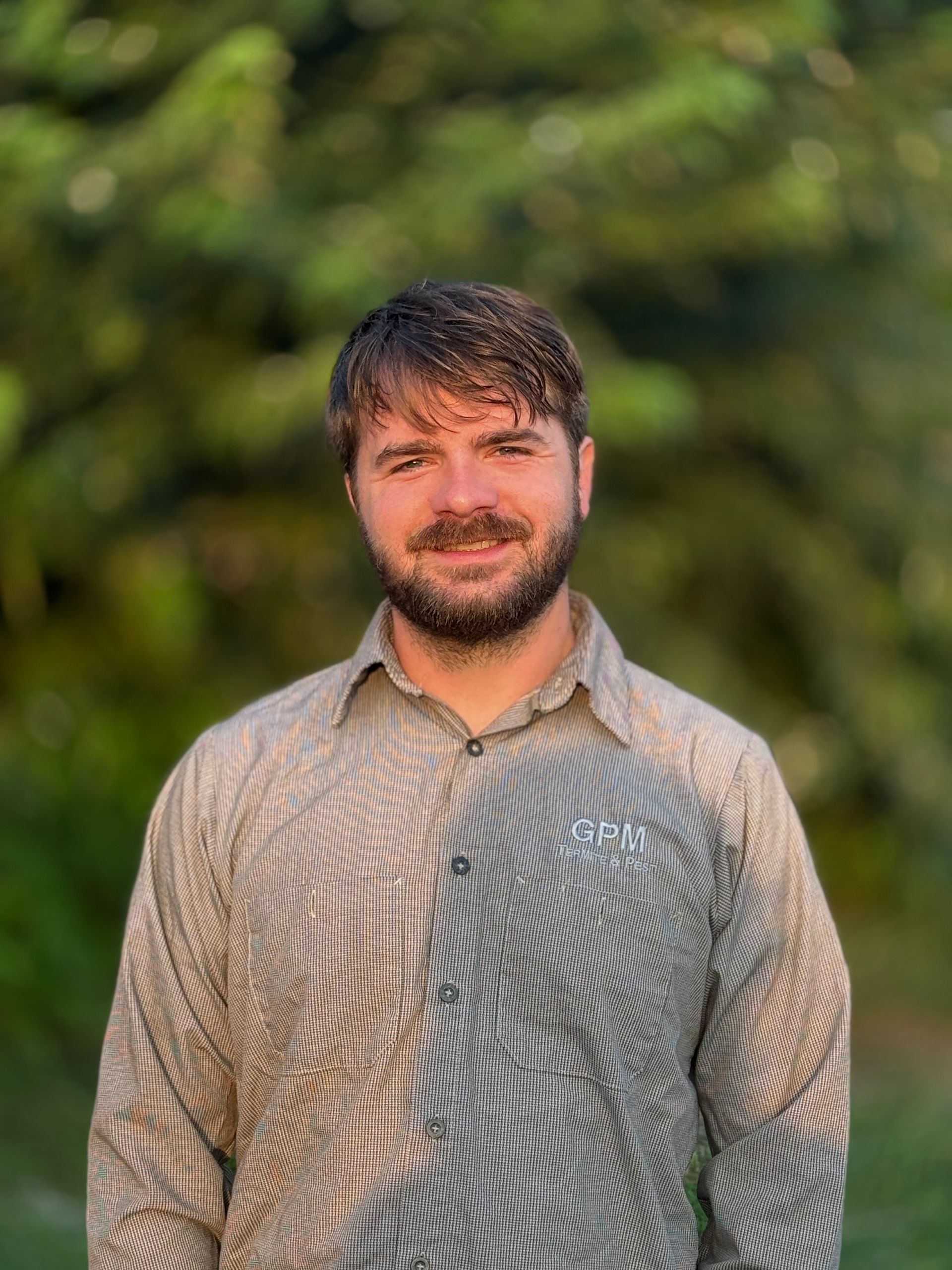A man with a beard and a striped shirt is standing in front of trees.