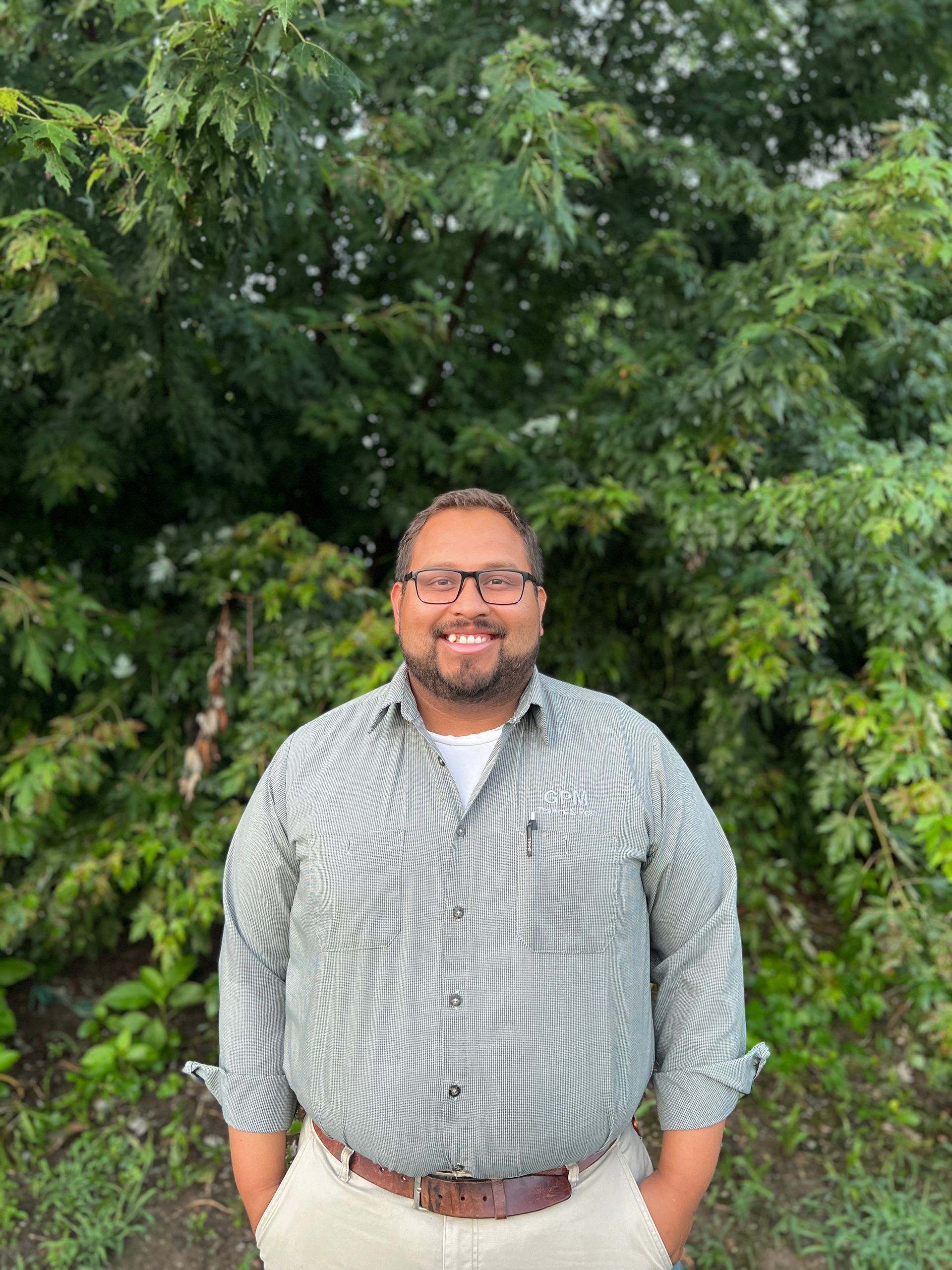 A man is standing in front of a forest with his hands in his pockets.