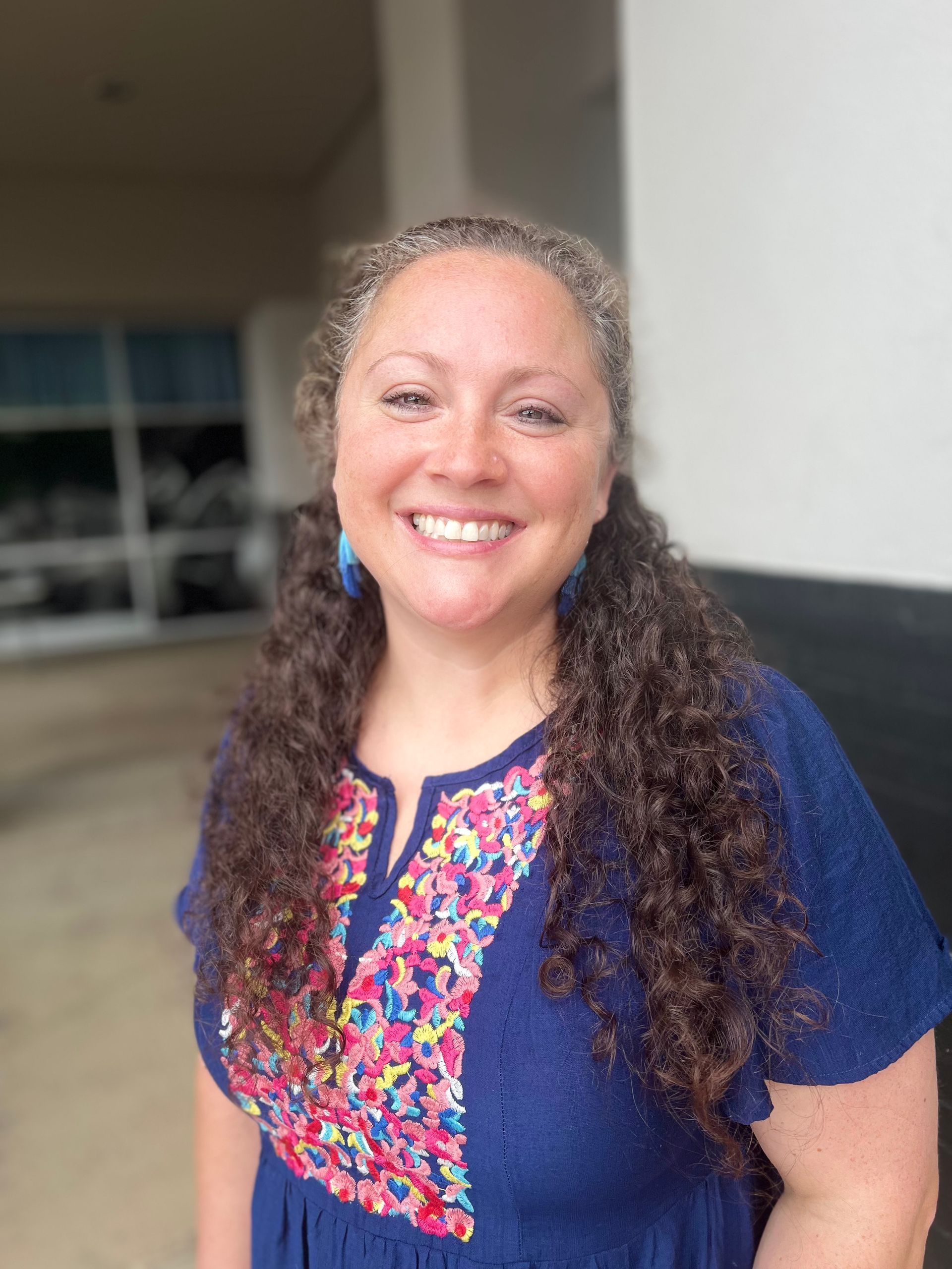 A woman with curly hair is wearing a blue dress and smiling.