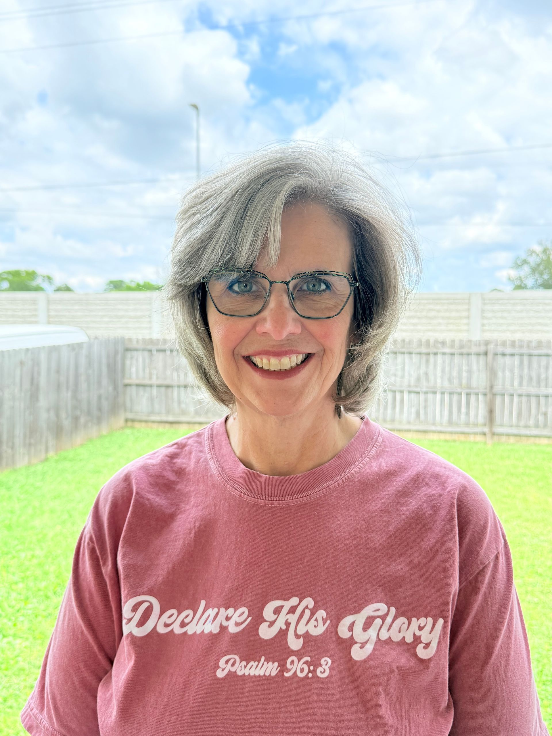A woman wearing glasses and a t-shirt that says `` declare his glory '' is smiling.