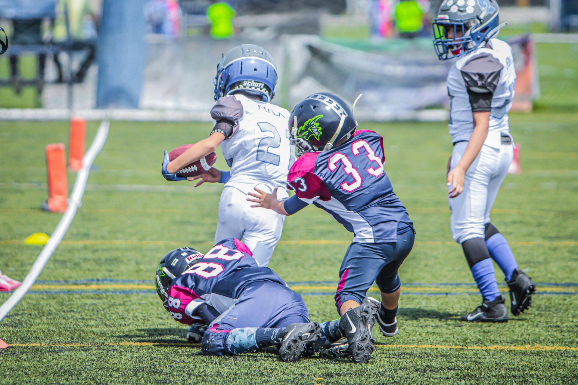 Children in football uniforms on a field, with one player tackled to the ground and another running with the ball.