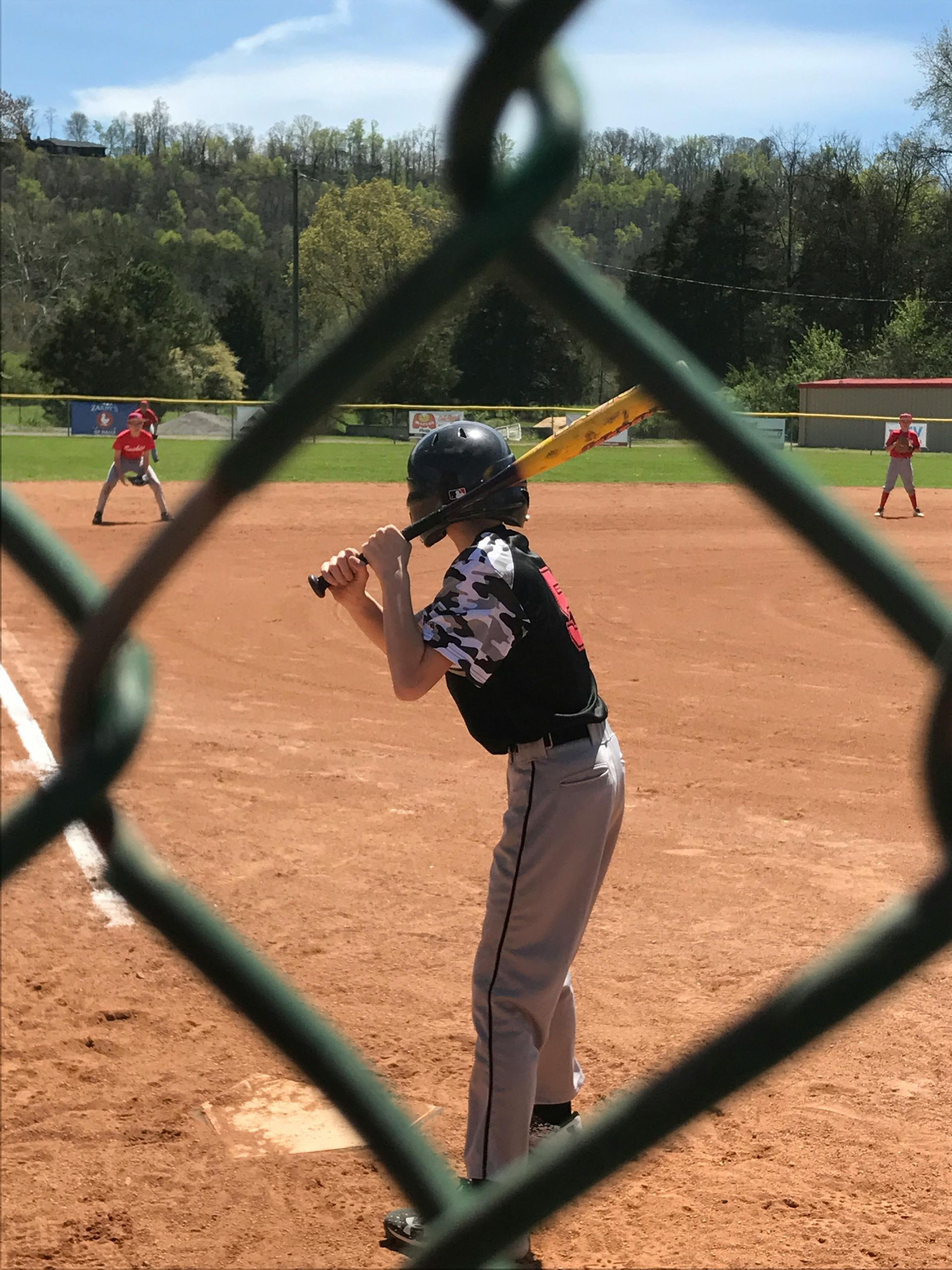 A baseball player in a black and patterned jersey stands at home plate, viewed through a chain-link fence.