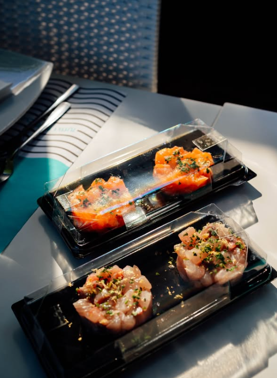 Due contenitori di plastica nera contengono salmone e tartare di pesce con erbe aromatiche.