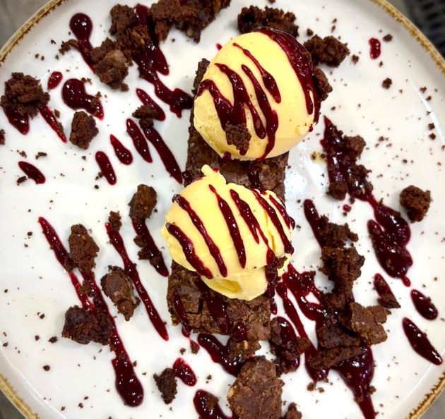 Dessert brownie con gelato alla vaniglia e salsa ai frutti di bosco su un piatto bianco.