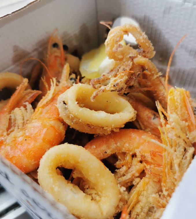 Frutti di mare fritti, tra cui gamberi e calamari, in una scatola di carta bianca con uno spicchio di limone.