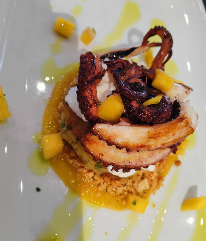 Polpo alla griglia con pezzi di mango su un letto di couscous e salsa gialla, piatto bianco.