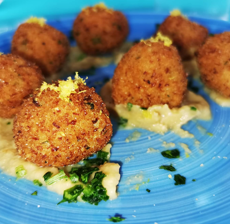 Palline dorate fritte su salsa cremosa, guarnite con erbe aromatiche e scorza di limone, servite su un piatto blu.