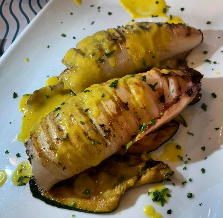 Calamari grigliati con salsa gialla su un piatto bianco, serviti con zucchine grigliate e un filo di prezzemolo.