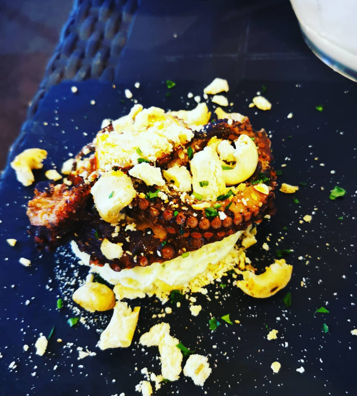 Piatto di polpo alla griglia con base bianca, guarnito con crostini ed erbe aromatiche su un piatto nero.