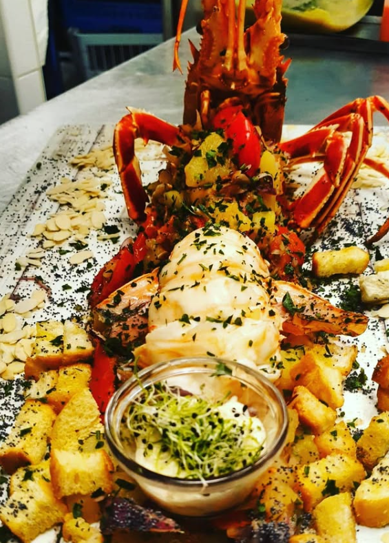 Piatto di aragosta con le chele distese, guarnito con erbe aromatiche e servito con una piccola ciotola di salsa e pane.