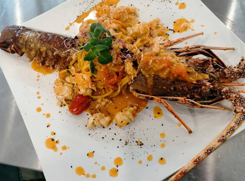 Piatto di aragosta su piatto bianco con pasta, pomodori, salsa ed erbe aromatiche.