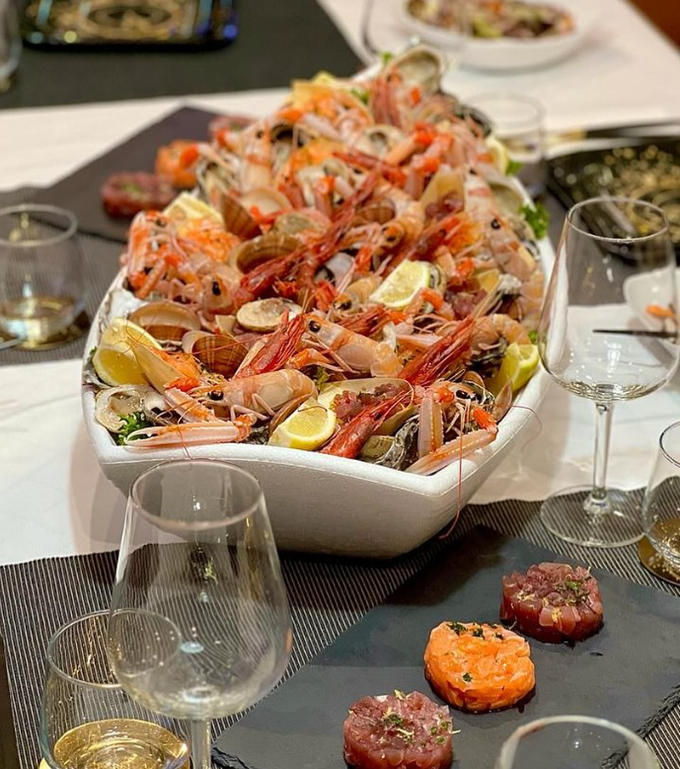 Piatto di frutti di mare con gamberi, ostriche e limone; quattro antipasti tartari su un tavolo con bicchieri da vino.