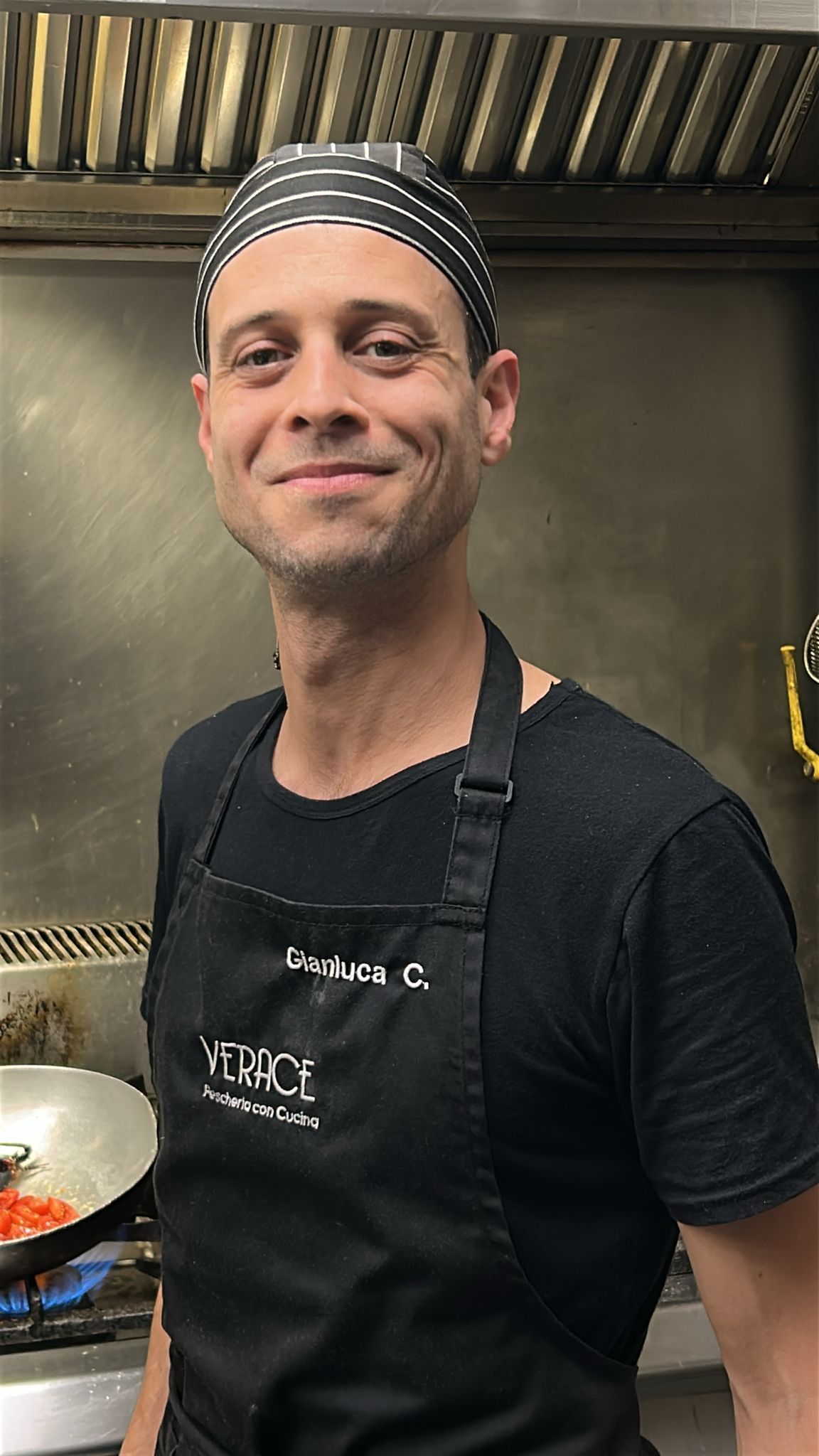 Chef in cucina con grembiule nero e cappello fantasia, sorridente.