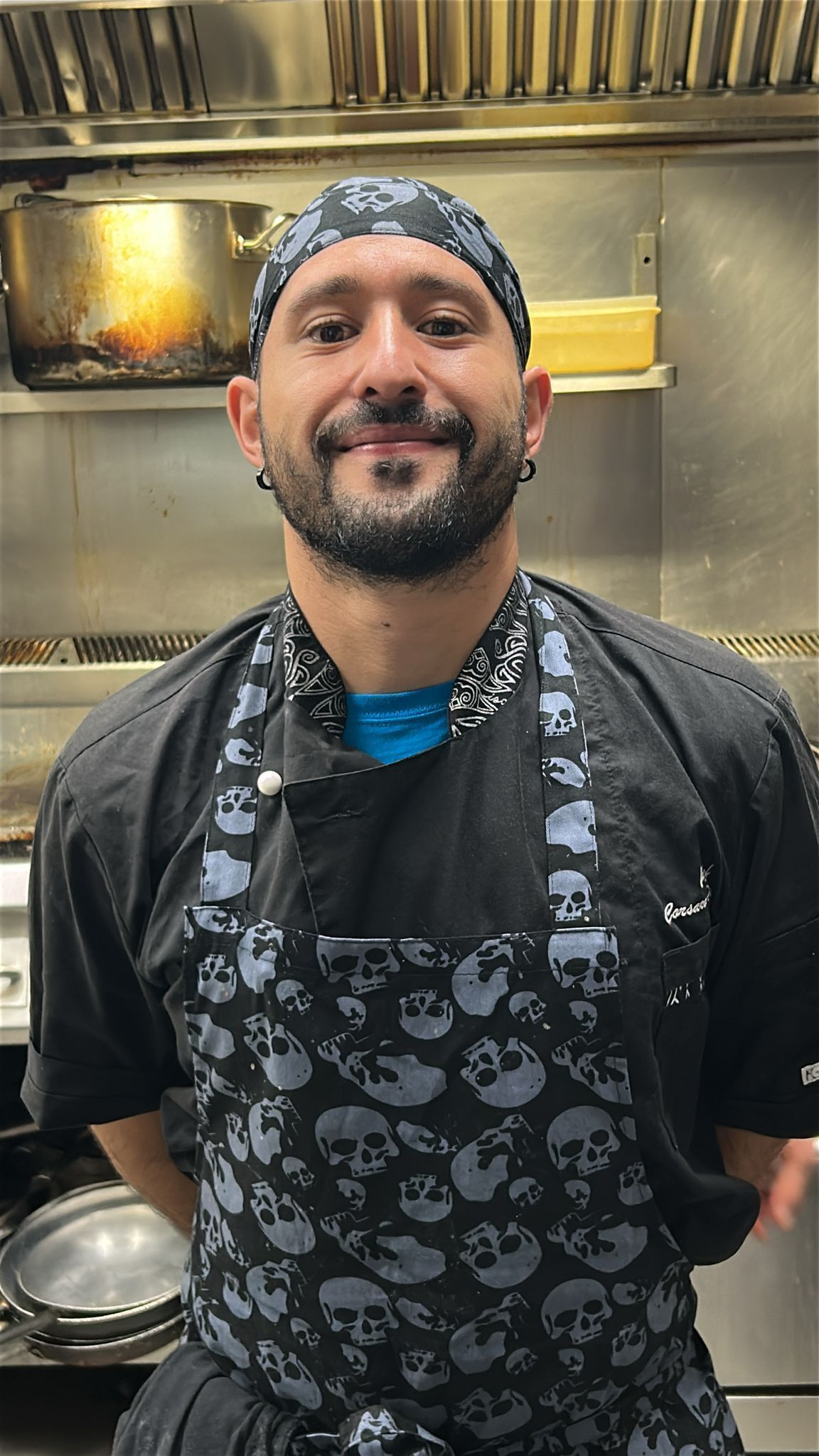 Cuoco sorridente in cucina, con indosso una bandana e un grembiule con un motivo a teschio.