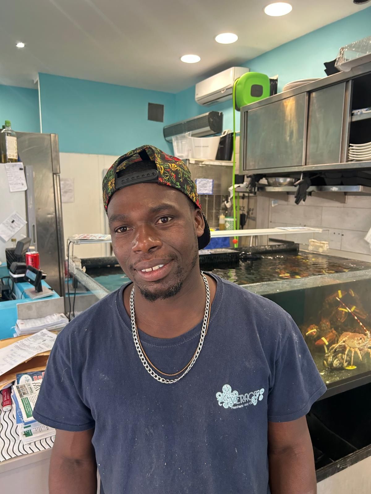 Un uomo con berretto e collana sorride in un negozio di pesce.