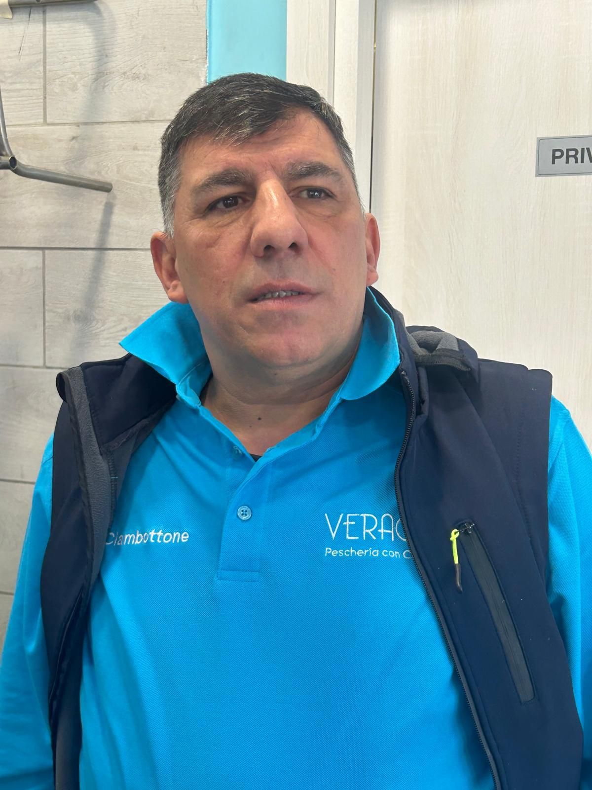 Uomo che indossa una polo blu e un gilet blu navy, guarda avanti con espressione neutra. Ambiente interno, cartello con la scritta