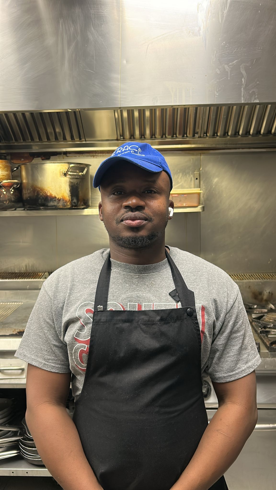 Un uomo con un berretto blu e un grembiule nero è in piedi in cucina e guarda la telecamera.