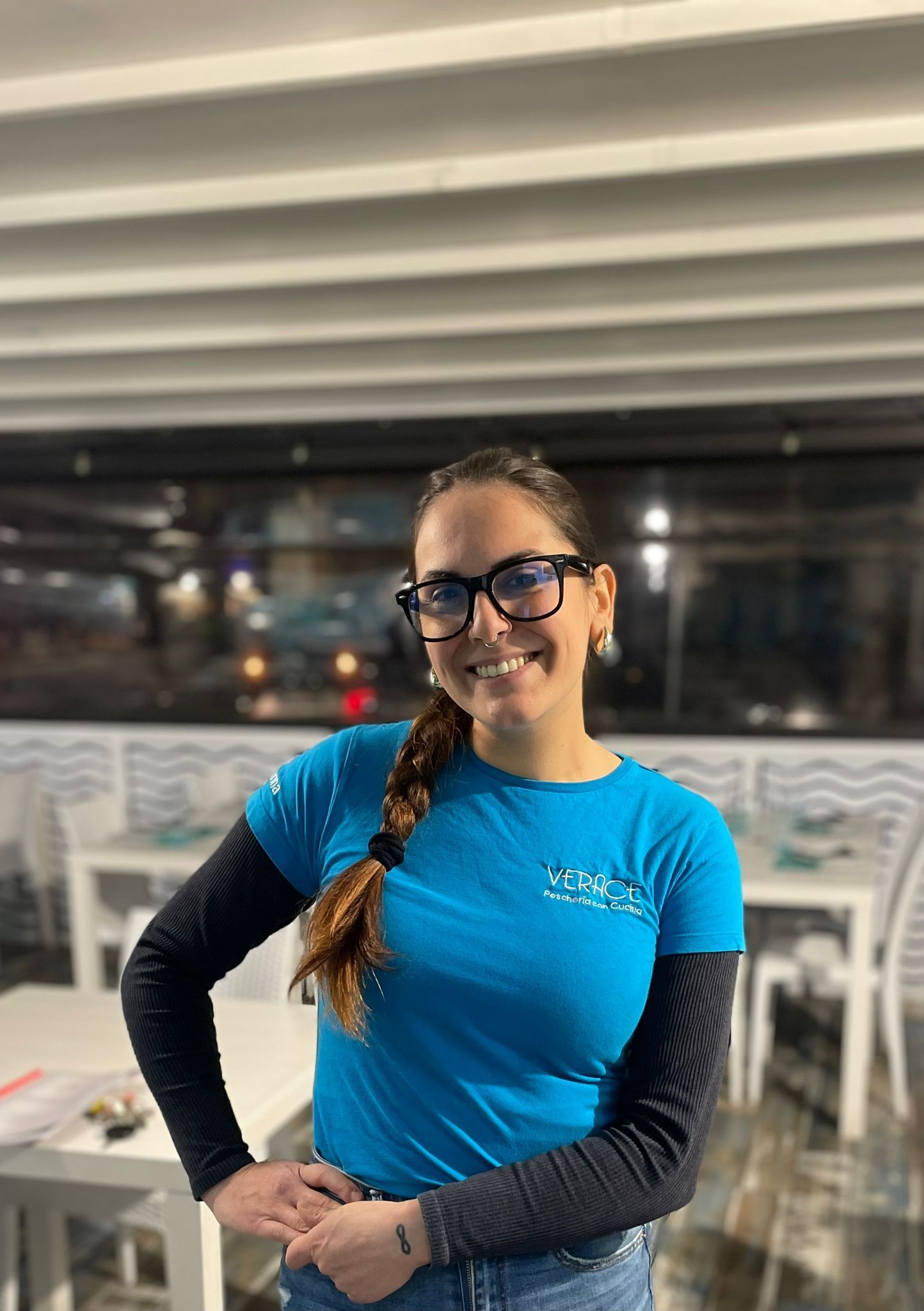 Donna con camicia blu, occhiali e treccia, sorridente con le mani sui fianchi, in un ristorante.