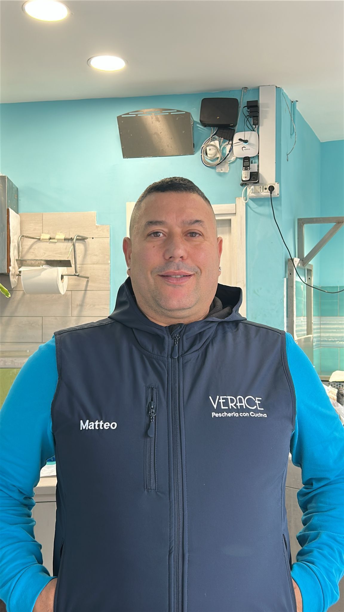 Un uomo che indossa un gilet blu navy e una camicia verde acqua sorride in un interno. Sullo sfondo, una parete verde acqua brillante.