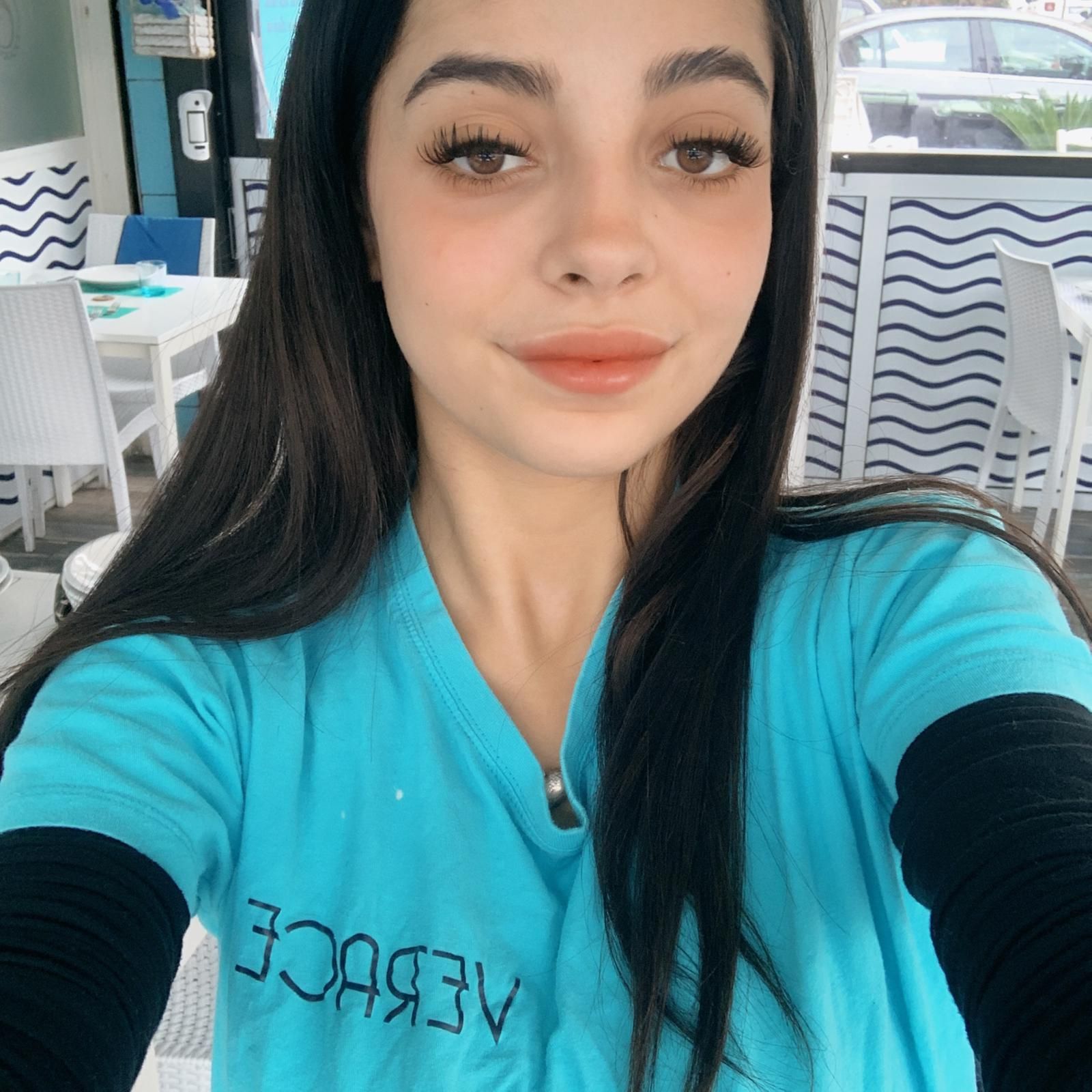 Donna con lunghi capelli scuri, che indossa una camicia turchese, sorridente, in un ristorante.