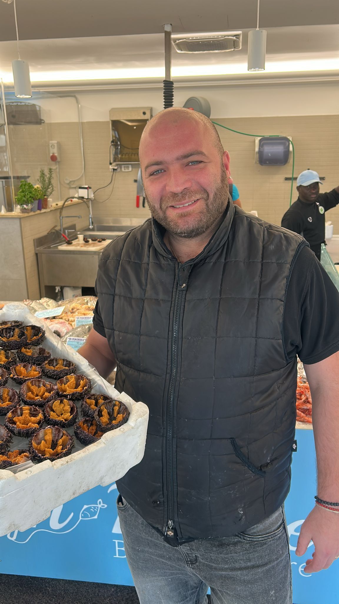 Un uomo sorride alla bancarella del mercato tenendo in mano un vassoio di cibo, probabilmente pesce.
