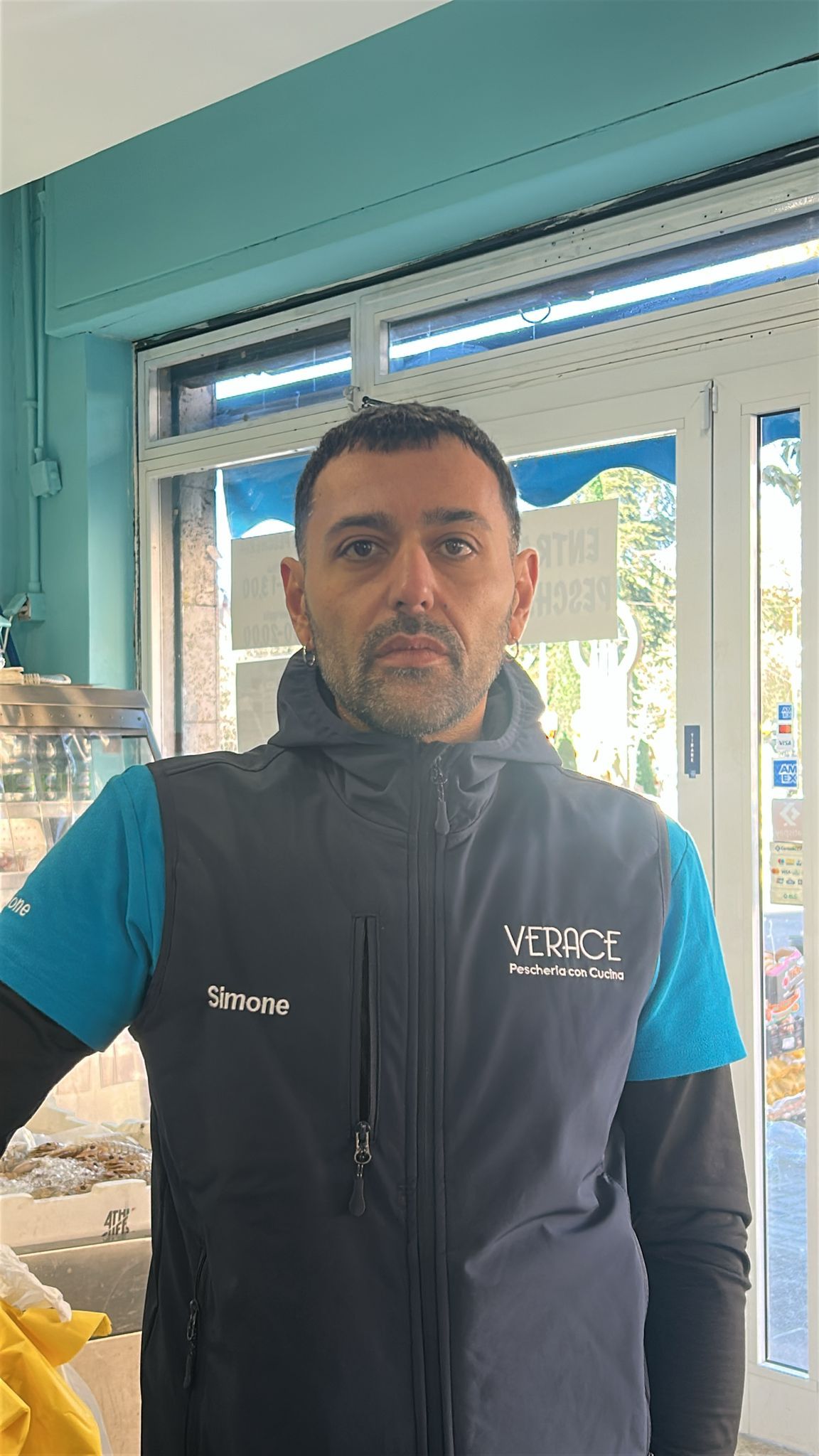 Uomo che indossa un gilet blu e nero, in piedi davanti a una porta a vetri.