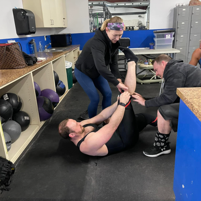 Coach assisting a male athlete with stretching exercises on the gym floor, focusing on proper technique and injury prevention.