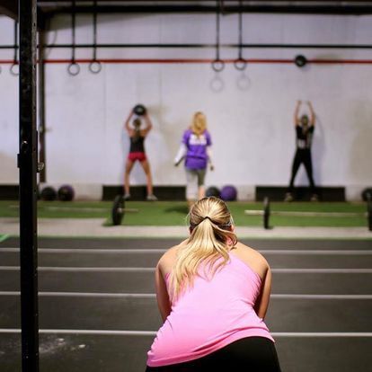 Woman in pink tank top observing a strength training class at a gym, focusing on participants lifting weights in the background.