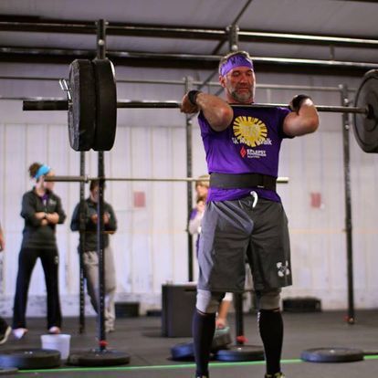 Mature male powerlifter in a purple Kansas Strength t-shirt lifting a heavy barbell at a gym competition, surrounded by spectators.