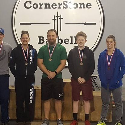Group of athletes with medals standing in front of a Barbell Club sign at Kansas Strength and Performance, celebrating competition success.