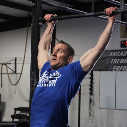 Middle-aged man wearing a Kansas Strength and Performance t-shirt while performing pull-ups in a gym, demonstrating upper body strength.