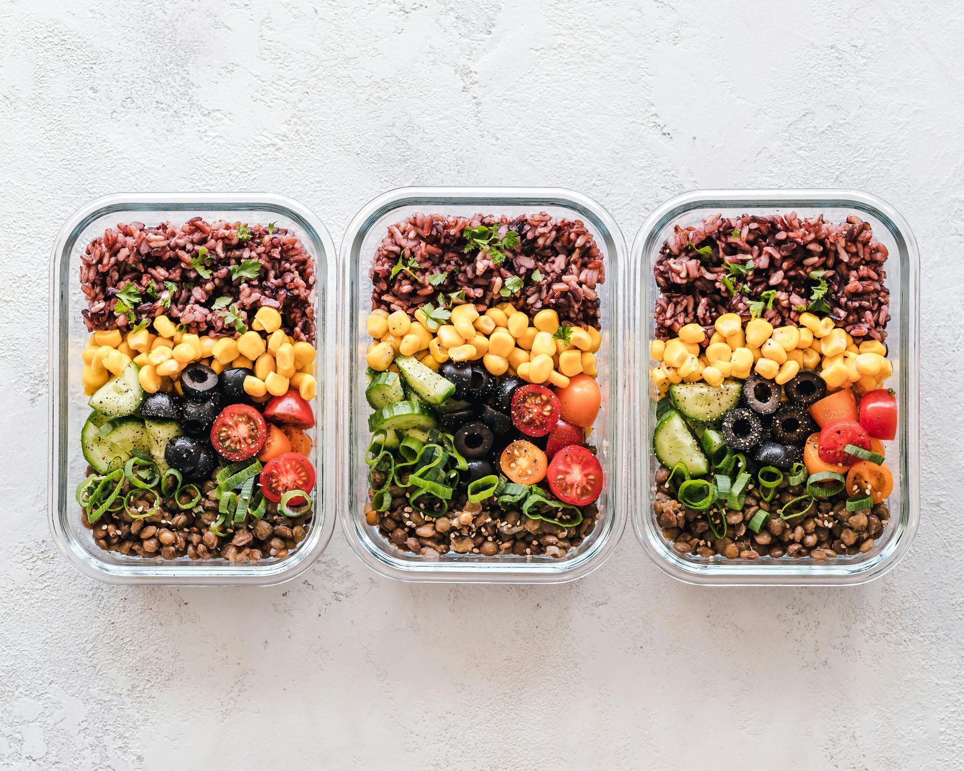 Three meal prep containers filled with brown rice, chickpeas, cucumbers, tomatoes, olives, and green peppers, arranged on a white surface, ideal for powerlifters’ nutrition.