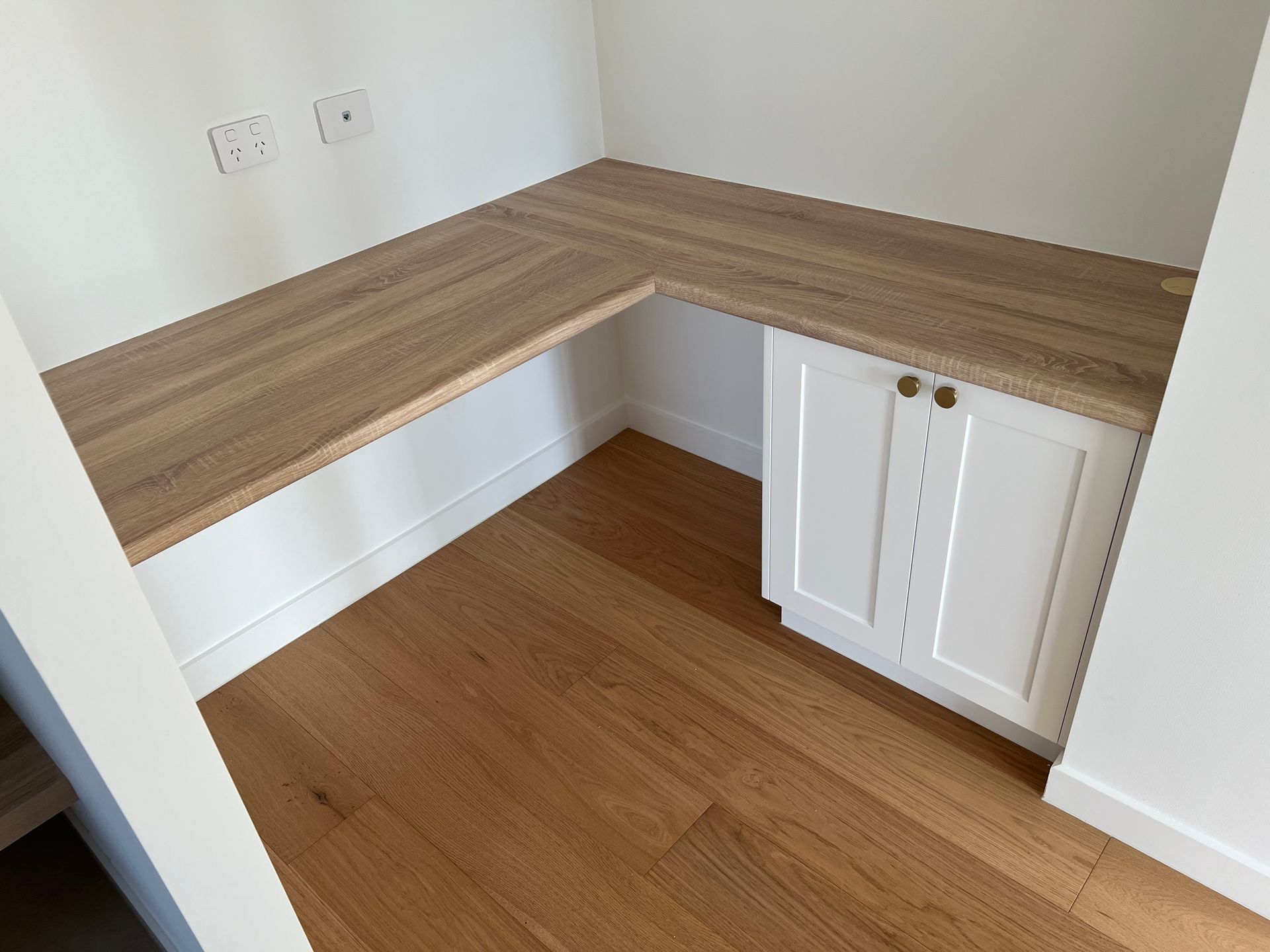 A corner of a room with a wooden desk and white cabinets.