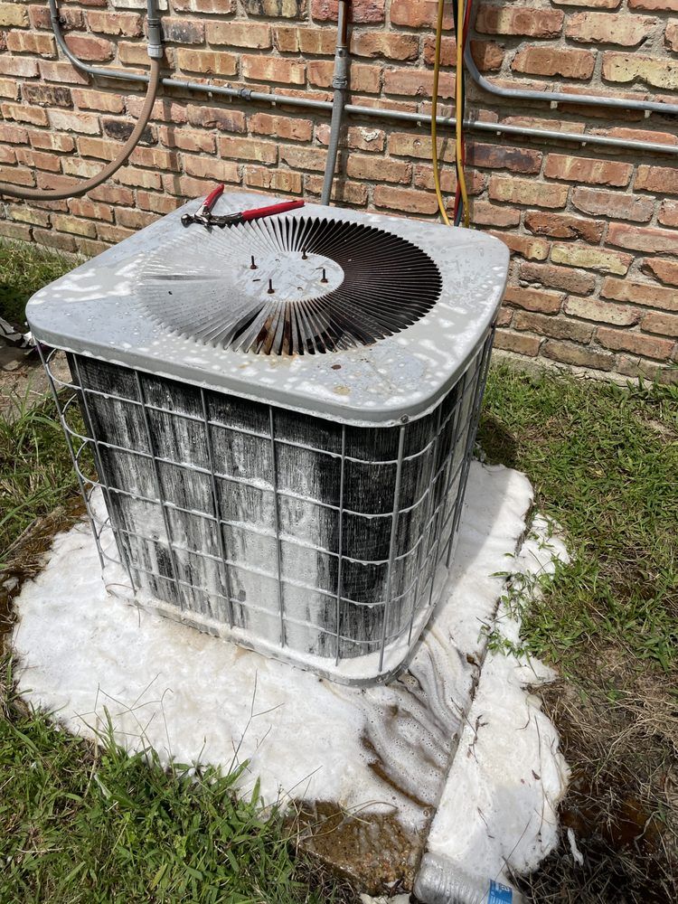 An outdoor air conditioning unit covered in thick, white cleaning foam sitting on a concrete pad against a brick wall.